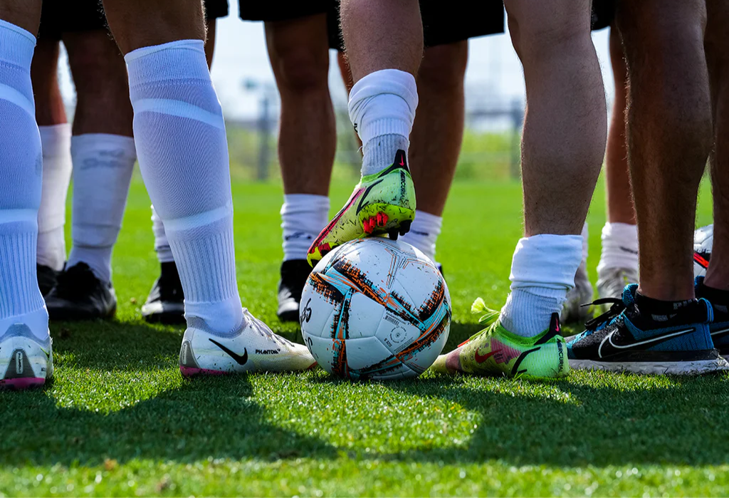 Senda Apex Match Soccer Ball surrounded by players' legs and cleats during pre-game action on a grass soccer field, capturing an in-play atmosphere.