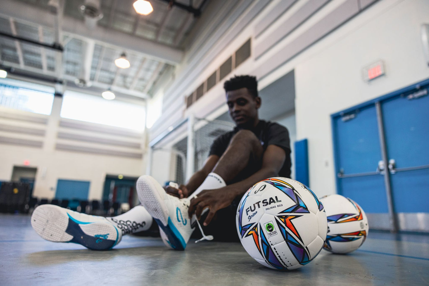 Indoor futsal player lacing up shoes beside Senda Palmas Eco Futsal Ball, showcasing its design and Fair Trade certification.