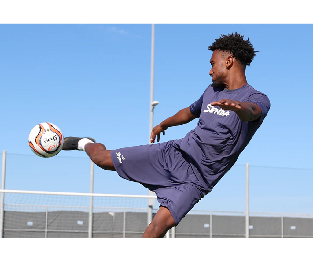 Athlete mid-air kicking Senda Valor Match Soccer Ball during outdoor soccer training on turf field.