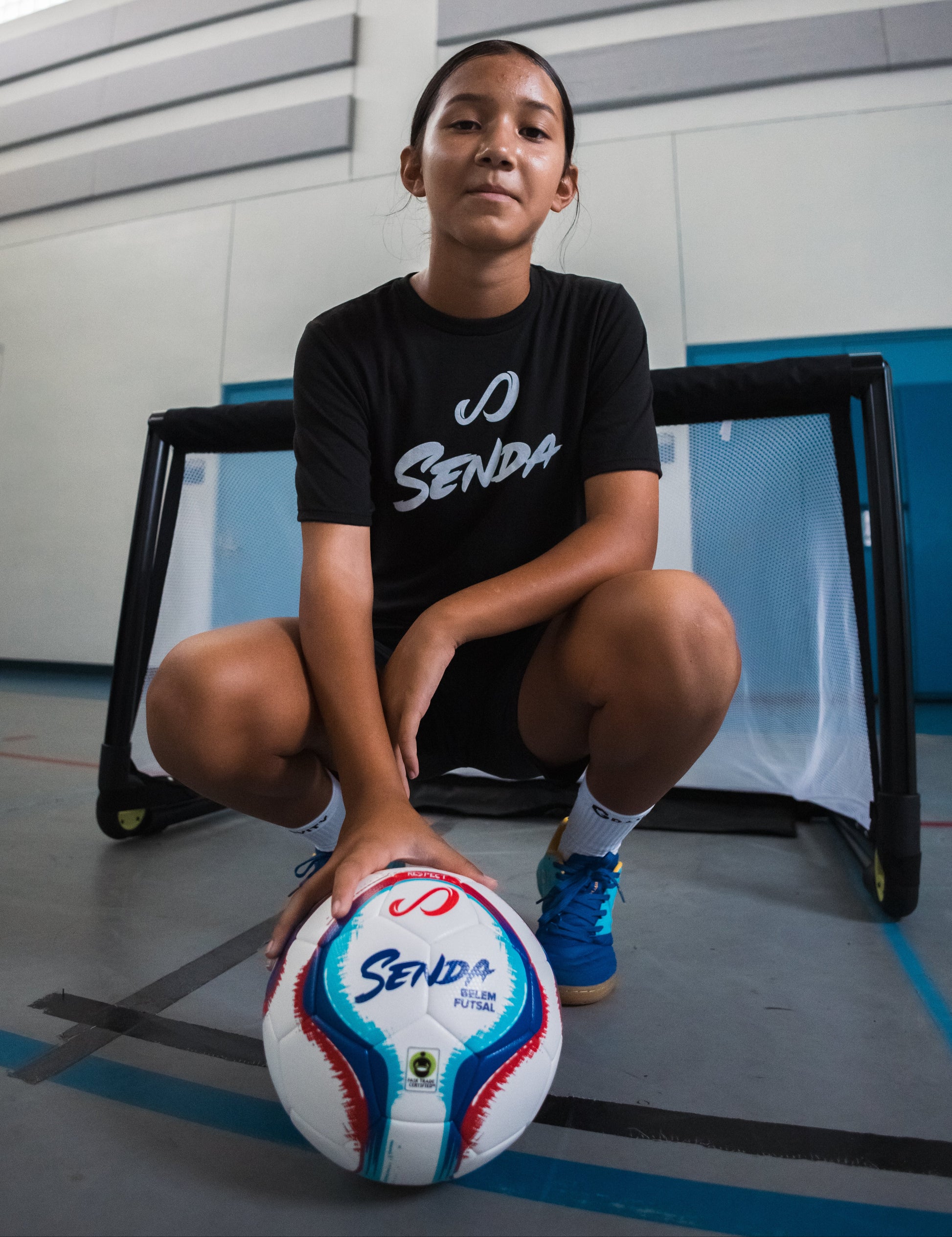 Young athlete posing with White Belem Training Futsal Ball by Senda – Fair Trade Certified futsal ball used in indoor training. Ideal for youth development and control-based play on hard courts.

