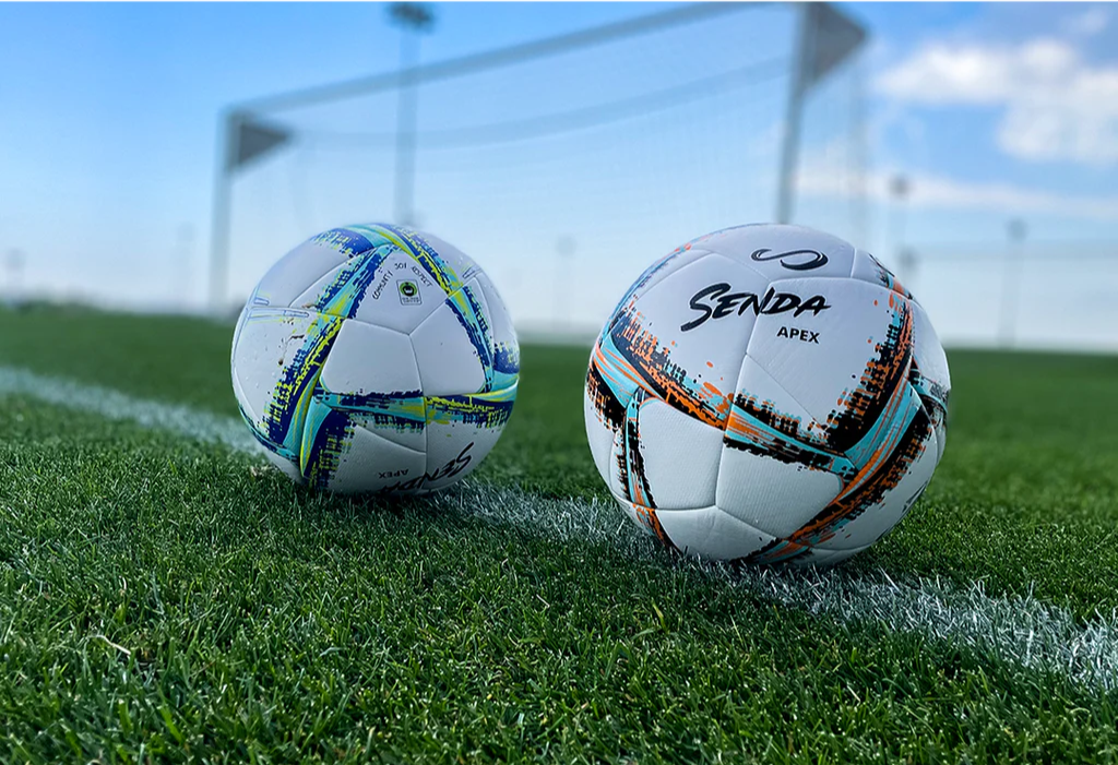 Senda Apex Match Soccer Balls in blue and orange variants resting on a grass soccer field with goalpost in background, showcasing real-play environment.







