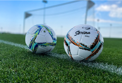 Senda Apex Match Soccer Balls in blue and orange variants resting on a grass soccer field with goalpost in background, showcasing real-play environment.







