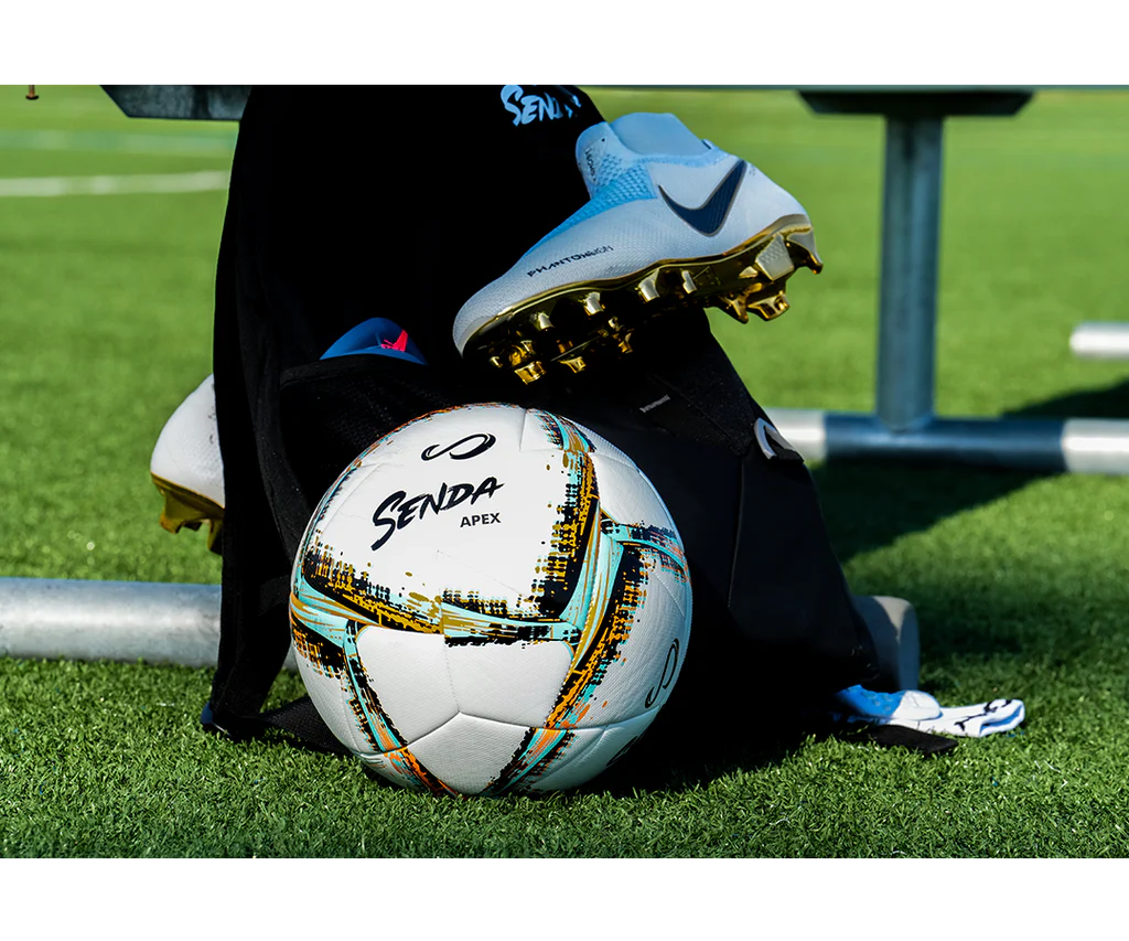 Senda Apex Match Soccer Ball placed beside Nike soccer cleats and training gear on a turf field, highlighting a real-game preparation scene.







