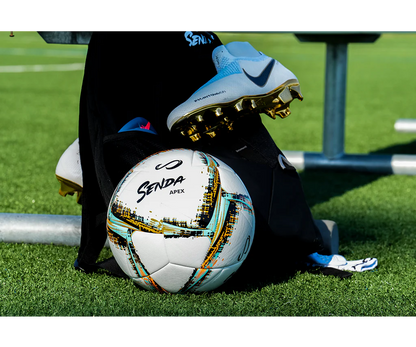 Senda Apex Match Soccer Ball placed beside Nike soccer cleats and training gear on a turf field, highlighting a real-game preparation scene.








