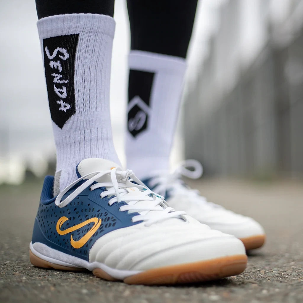 Side view of Senda white athletic socks with black branding, styled with white and navy futsal shoes on an outdoor court for a sporty street-ready look.

