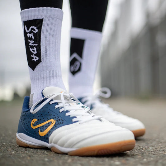 Side view of Senda white athletic socks with black branding, styled with white and navy futsal shoes on an outdoor court for a sporty street-ready look.

