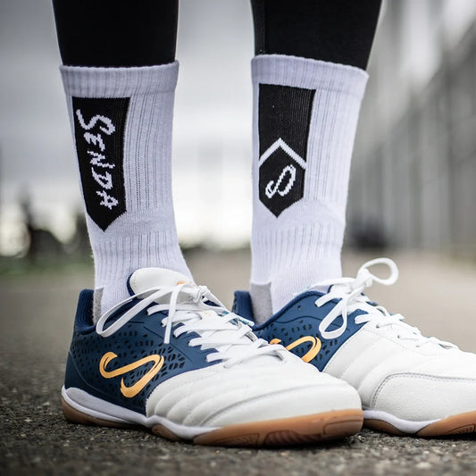 Close-up of Senda Athletic Socks in white with black logo design, worn with indoor soccer shoes on concrete surface, highlighting cushioned athletic support.

