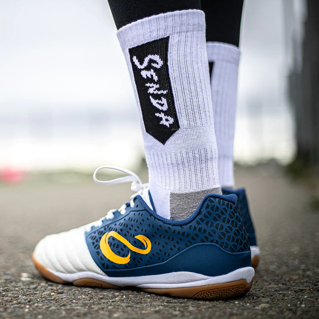 Side close-up of Senda white athletic socks with black logo, paired with navy and white futsal shoes on an outdoor surface, showing fit and texture.

