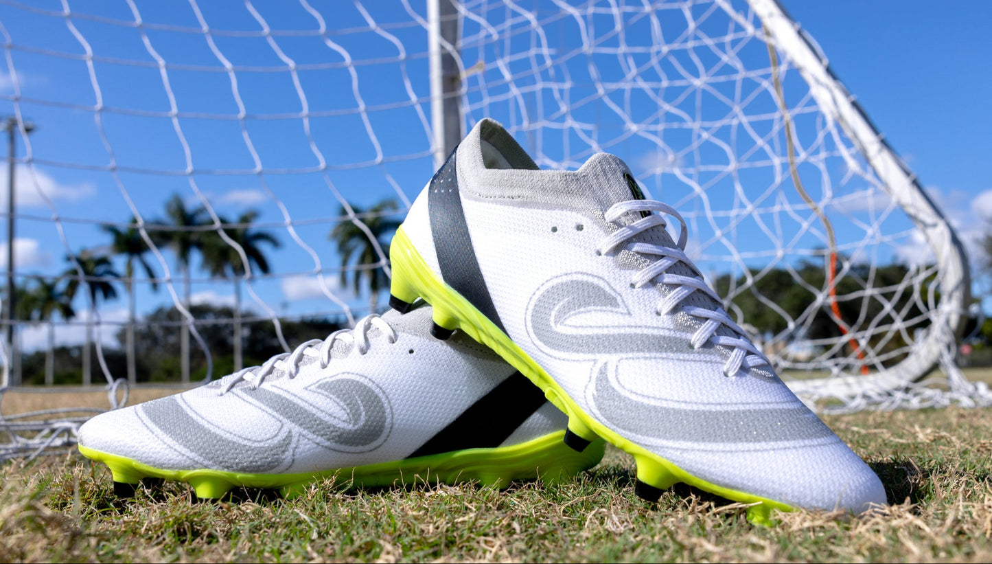 Pair of Senda Pampa Pro Soccer Cleats placed on grass field in front of goal net, highlighting aerodynamic design and neon stud outsole.

