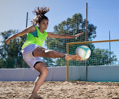 Female athlete barefoot kicking the Senda Playa Beach Soccer Ball on sand during a beach soccer game.


