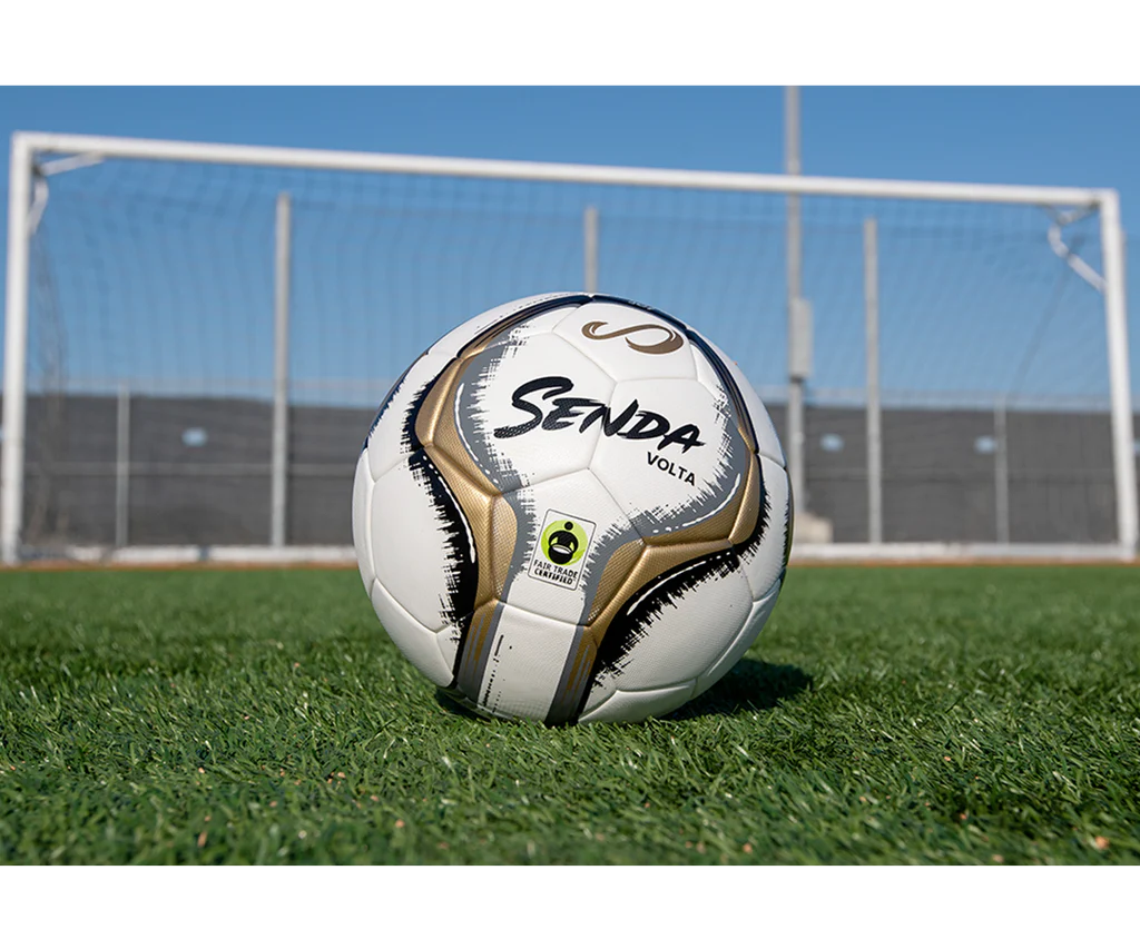 Senda Volta Professional Soccer Ball resting on green turf in front of a goal post under clear blue sky, highlighting game-ready design and Fair Trade label.

