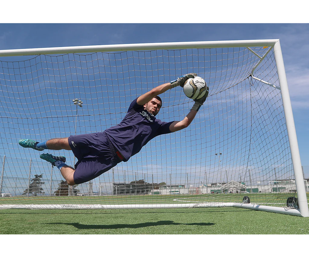 Goalkeeper diving mid-air to save a shot using the Senda Volta Professional Soccer Ball on a sunny field, showcasing ball control and match-level performance.

